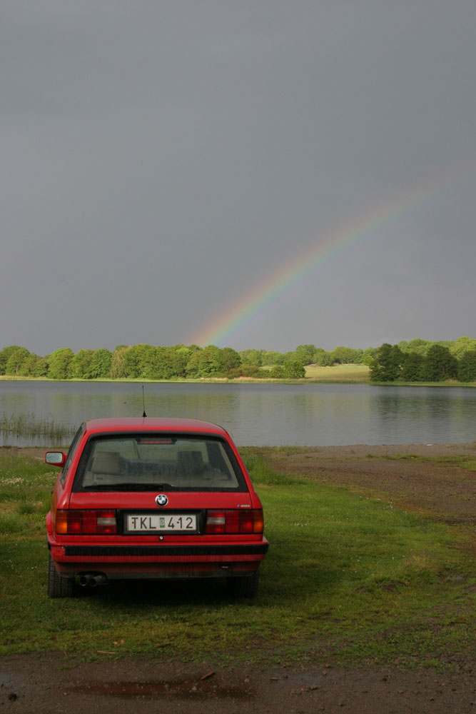 Röd BMW E30 bakifrån med regnbåge och sjö i bakgrunden