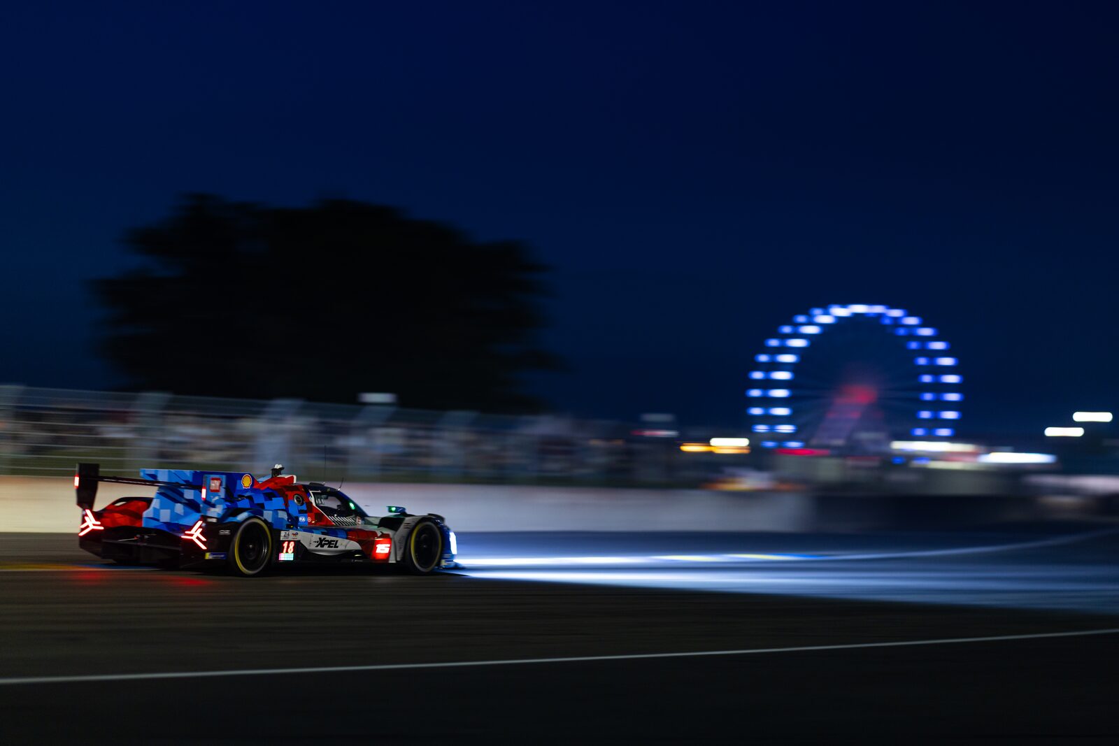 #15 BMW M Hybrid V8 i blå-röd kamouflagefärg i natten med Le Mans pariserhjul i bakgrunden