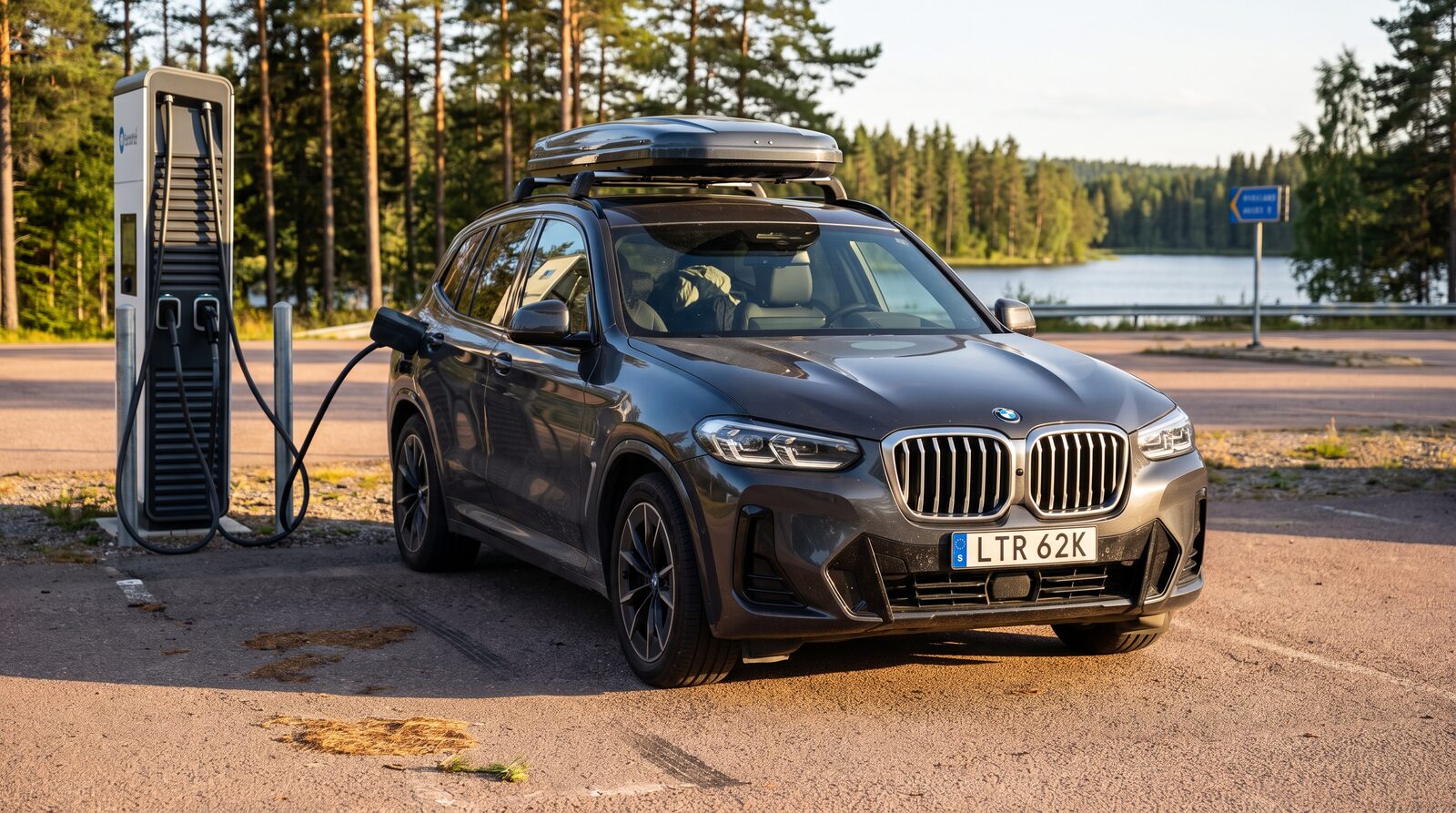 En mörkgrå BMW-elbil med takbox laddar vid en svensk motorvägsrastplats i sommarkvällsljus, med tallskog och sjö i bakgrunden.
