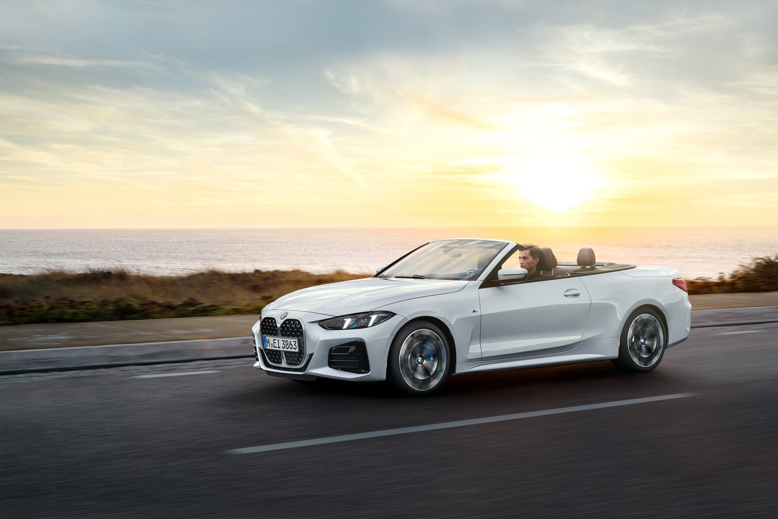 Vit BMW 4-serie Cabriolet i fart på kustväg i solnedgång