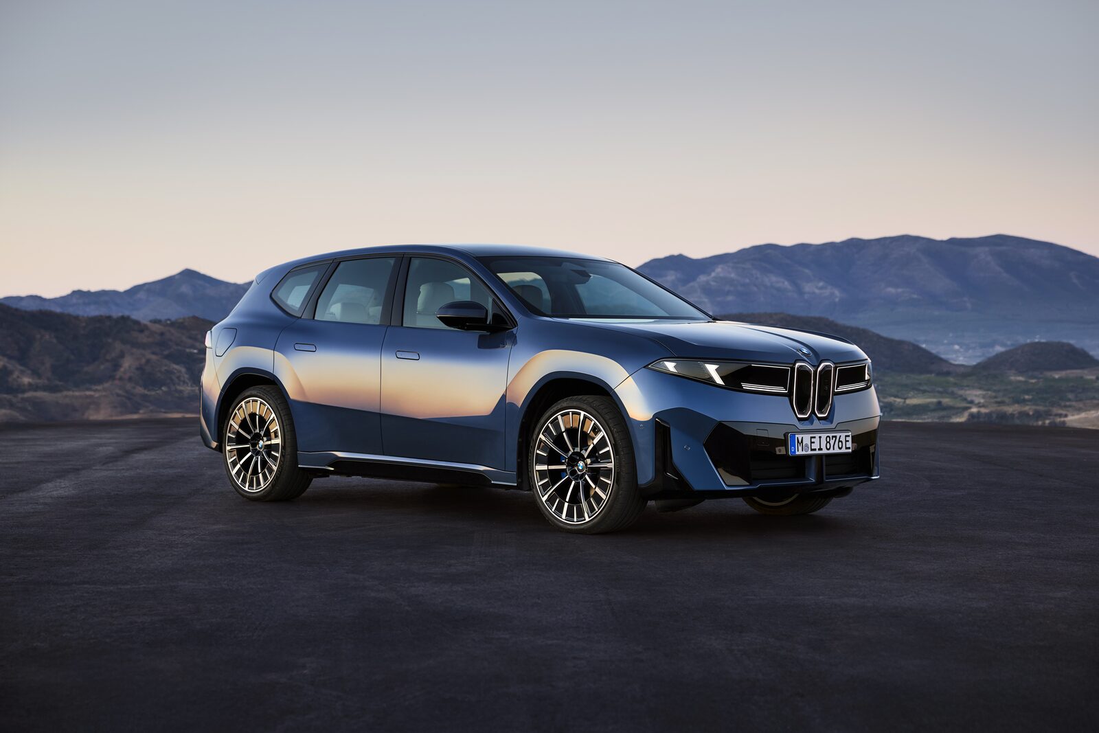 Blå BMW iX3 i trekvartsvy framifrån på grusplan med bergskedja i bakgrunden