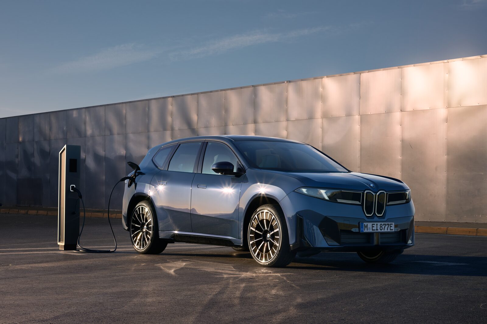 Blå BMW iX3 ansluten till en laddstolpe framför en metallfasad i skymningsljus