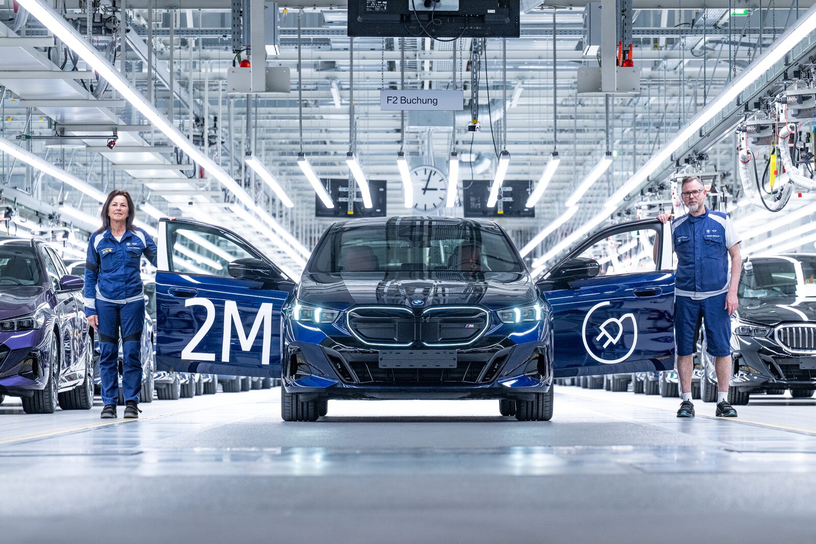Blå BMW i5 M60 xDrive på monteringslinan i Dingolfing mellan två uppfällda dörrar märkta 2M, flankerad av två fabriksarbetare