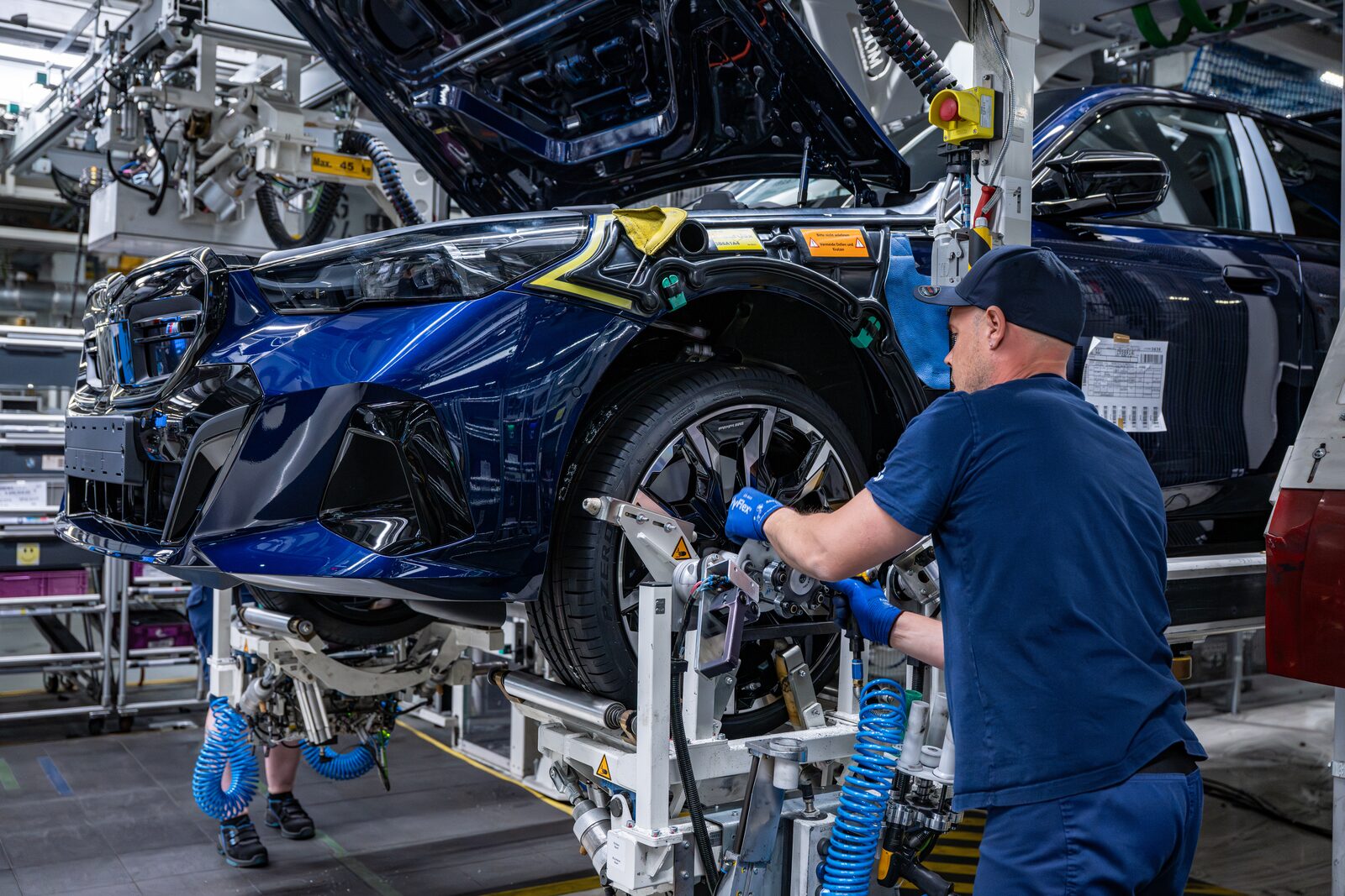 Fabriksarbetare i blå overall monterar en detalj vid framvagnen på en blå BMW i5 på linan