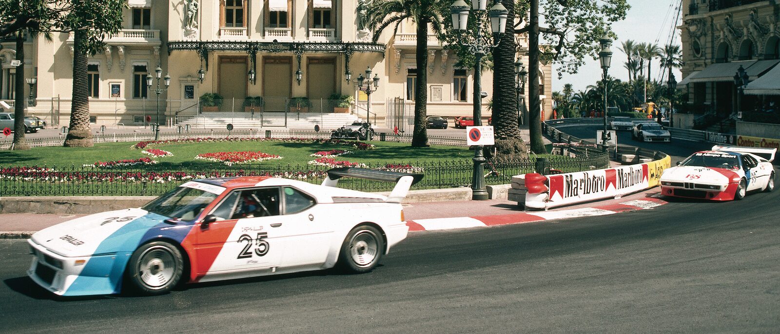 Två BMW M1 Procar i vit och blå lack på Monacos gator med kasinot och stadens fasader i bakgrunden