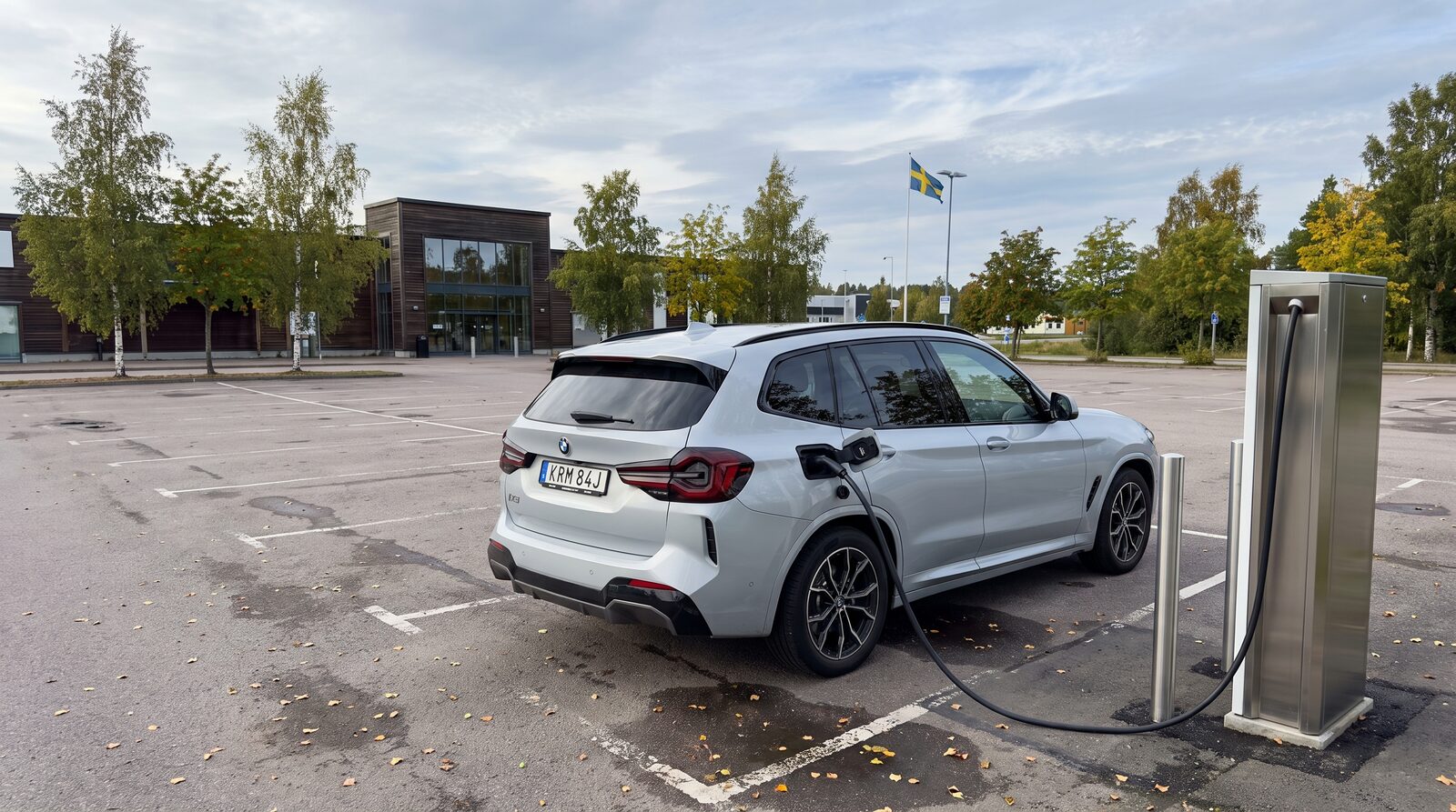 Vit BMW iX3 laddar vid en snabbladdare på en svensk parkering i sensommarljus