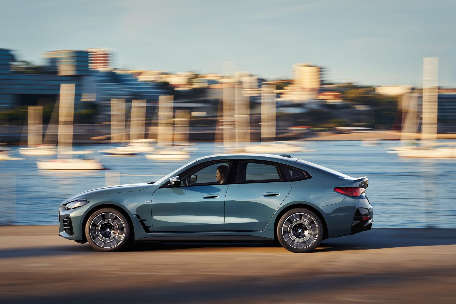 Grågrön BMW i4 i fart längs en kaj med suddig stadssiluett i bakgrunden