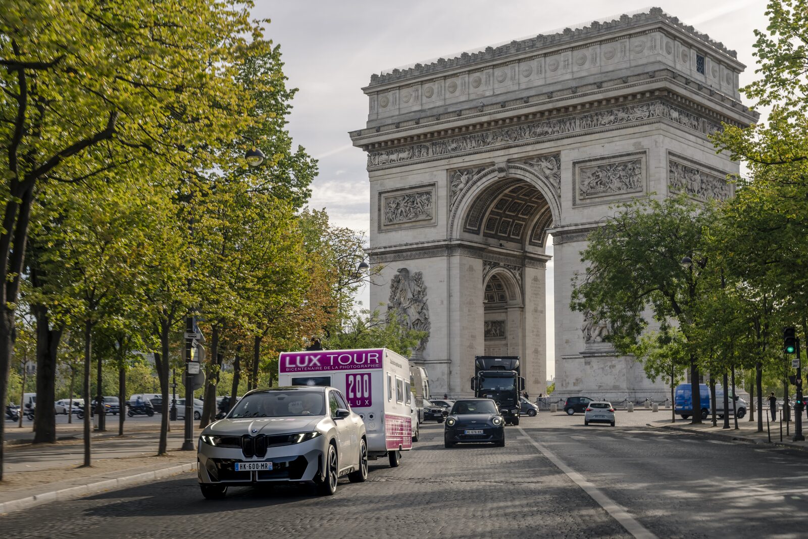 Silvergrå BMW iX3 drar en vit studiovagn med LUX TOUR-skylt på en allé i Paris med Triumfbågen i bakgrunden