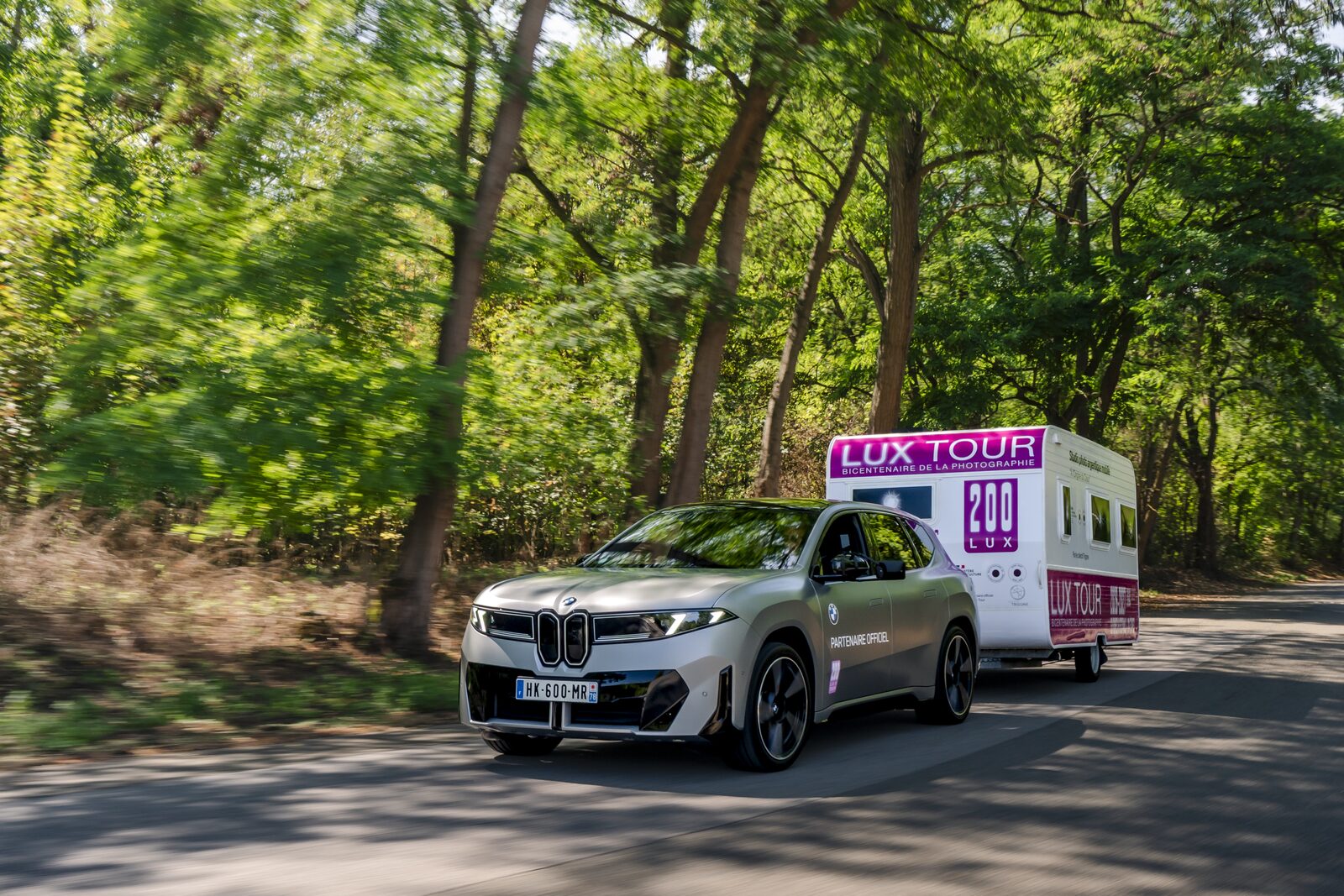 BMW iX3 med studiovagn i rörelse längs en trädkantad allé, fotograferad med panorering så bakgrunden är oskarp