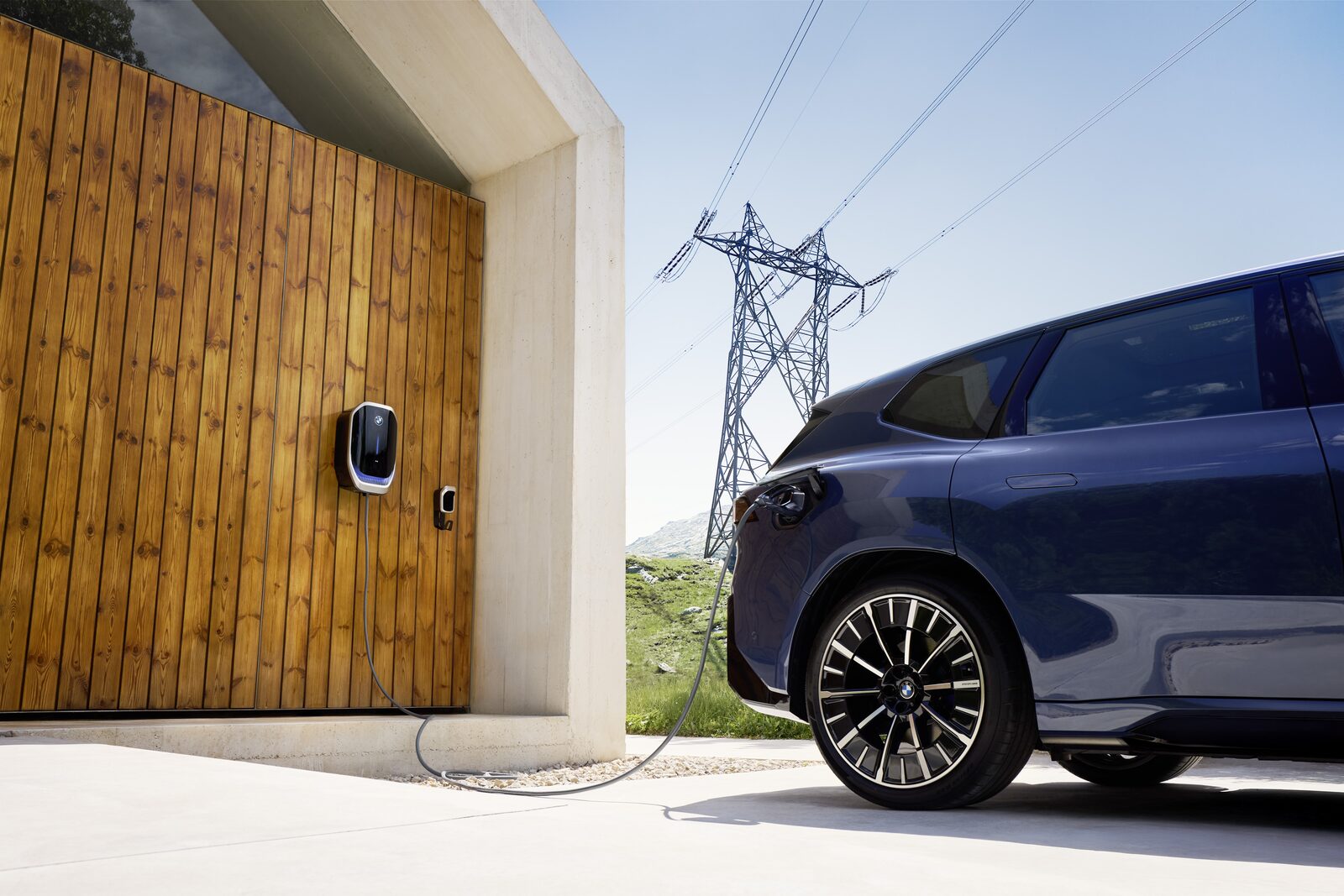 Mörkblå BMW iX3 laddar vid en BMW Wallbox monterad på en garageport i trä, med en kraftledningsstolpe i bakgrunden