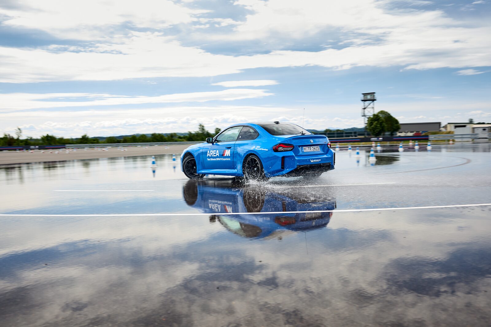 Blå BMW M2 i AREA M-dekor följer driftcirkeln på en blöt halkbana med vattenstänk från bakhjulen
