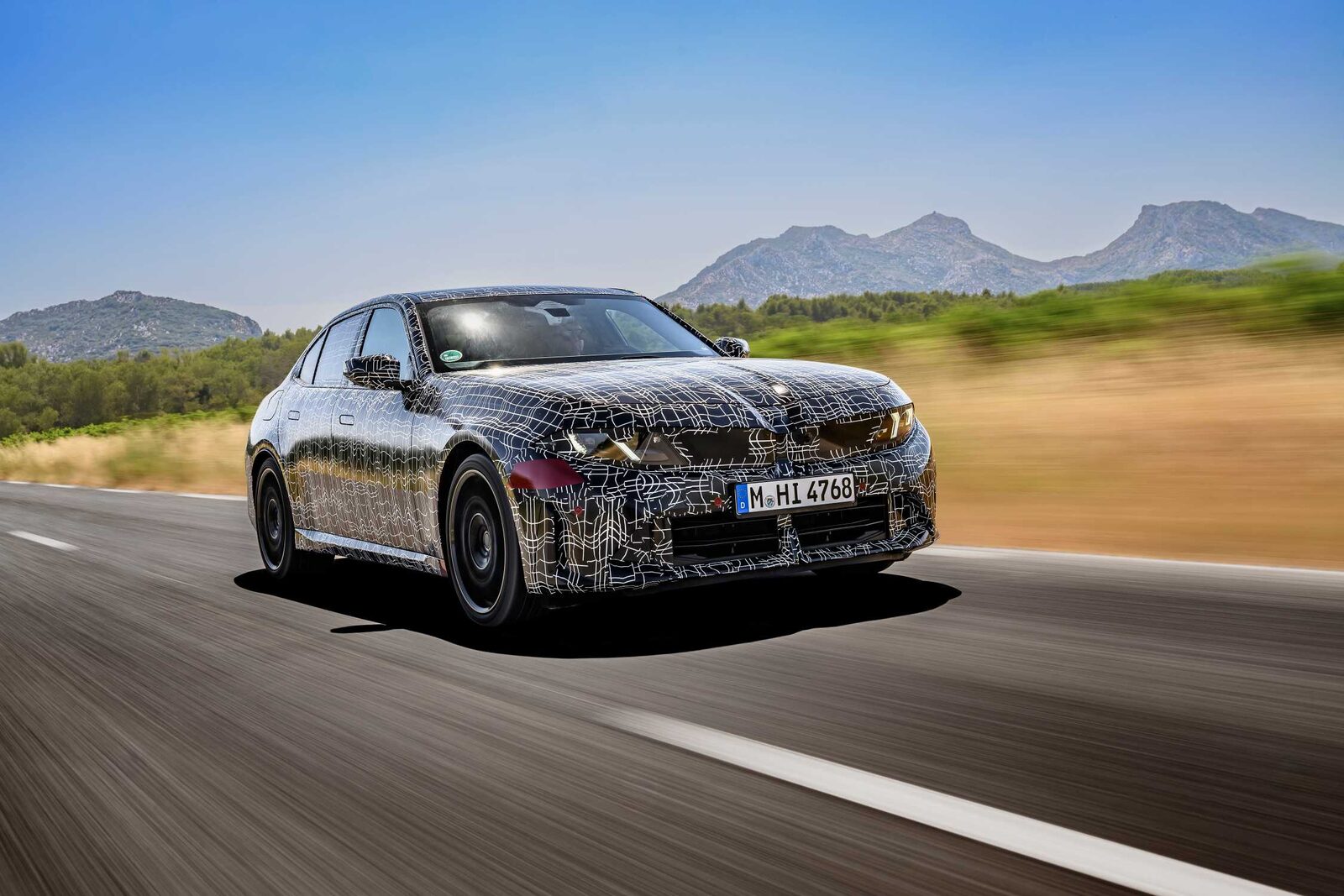 Kamouflerad prototyp av nya BMW 3-serie kör på landsväg i södra Frankrike med berg i bakgrunden
