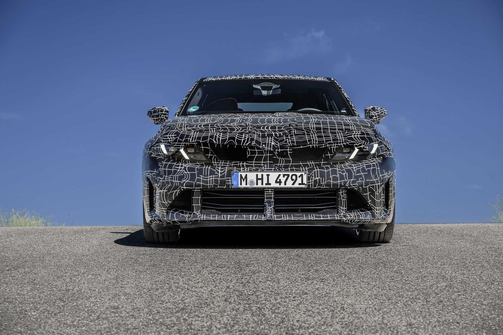 Kamouflerad BMW 3-serie-prototyp rakt framifrån mot blå himmel med de nya smala strålkastarna synliga