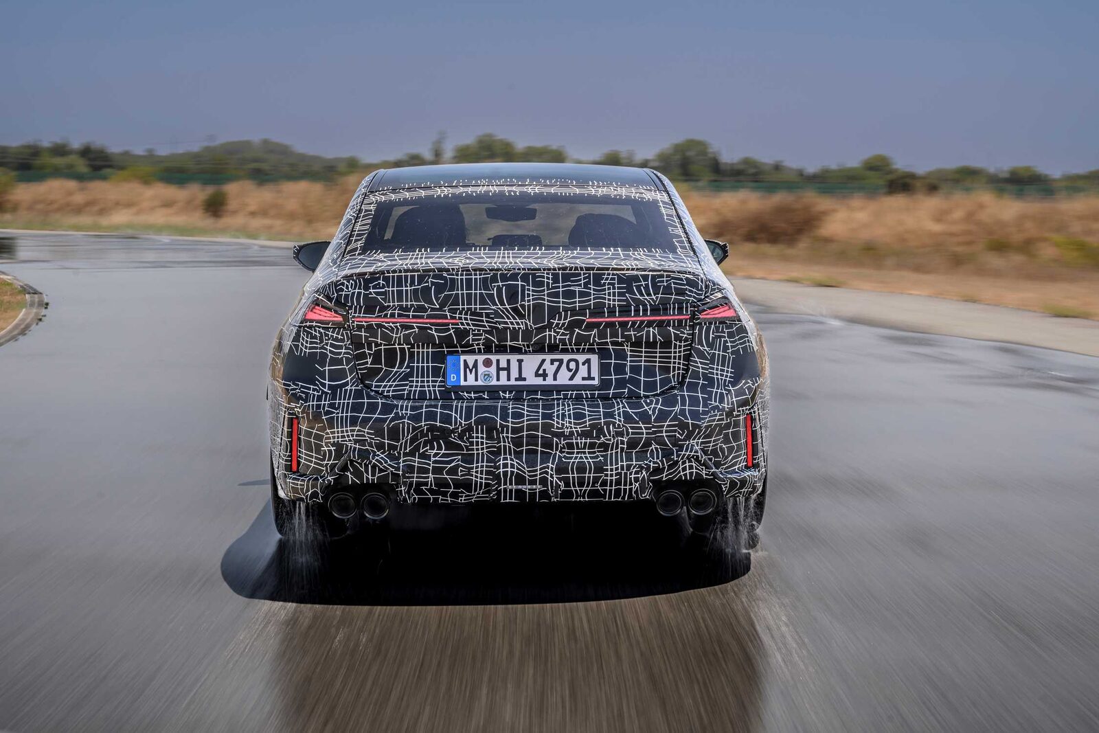 Kamouflerad BMW 3-serie-prototyp bakifrån på blöt asfalt med vattenspray från bakhjulen