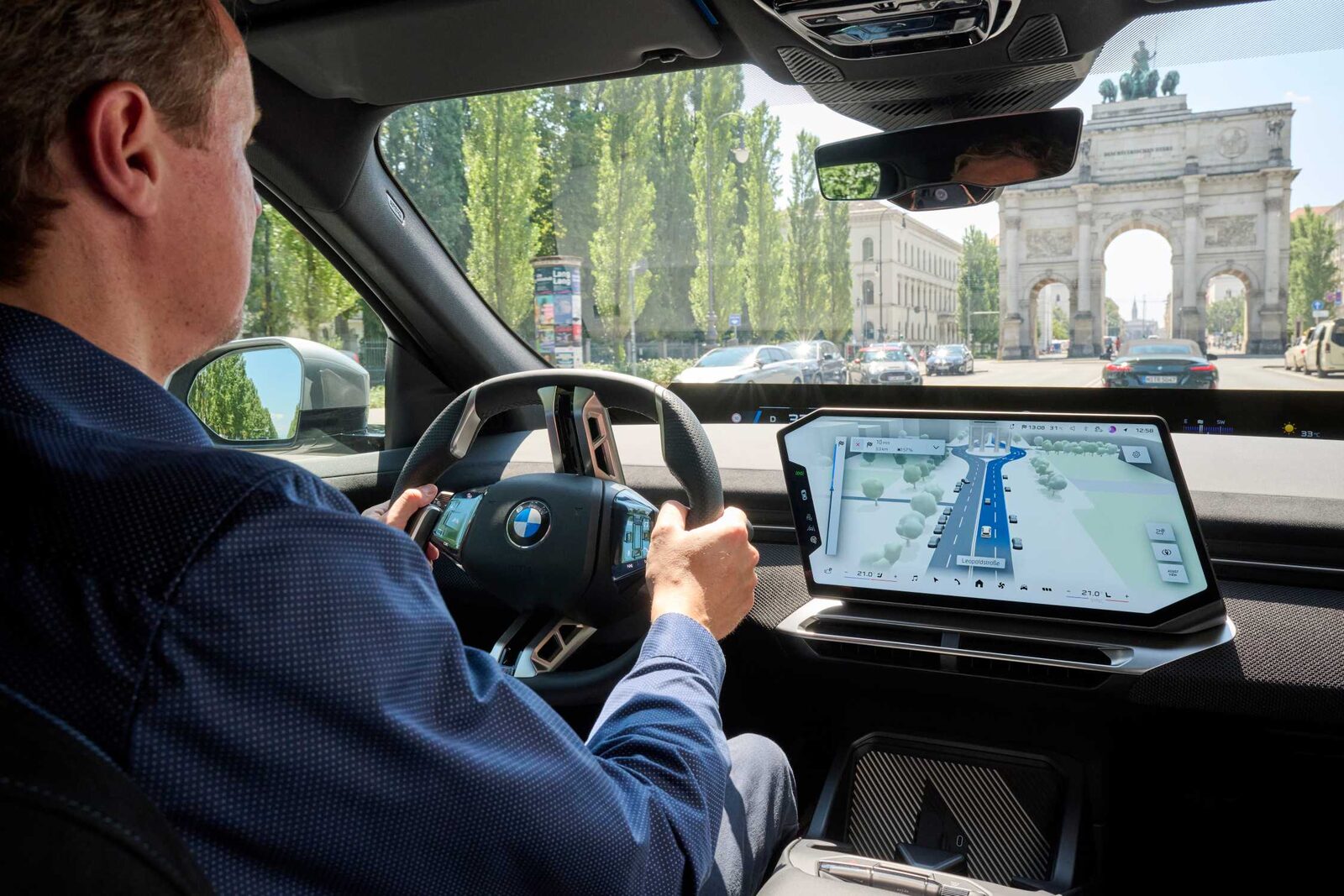 Förare i stadstrafik på Leopoldstraße i München med Siegestor framför bilen, mittskärmen visar en 3D-vy av gatan med träd och parkerade bilar