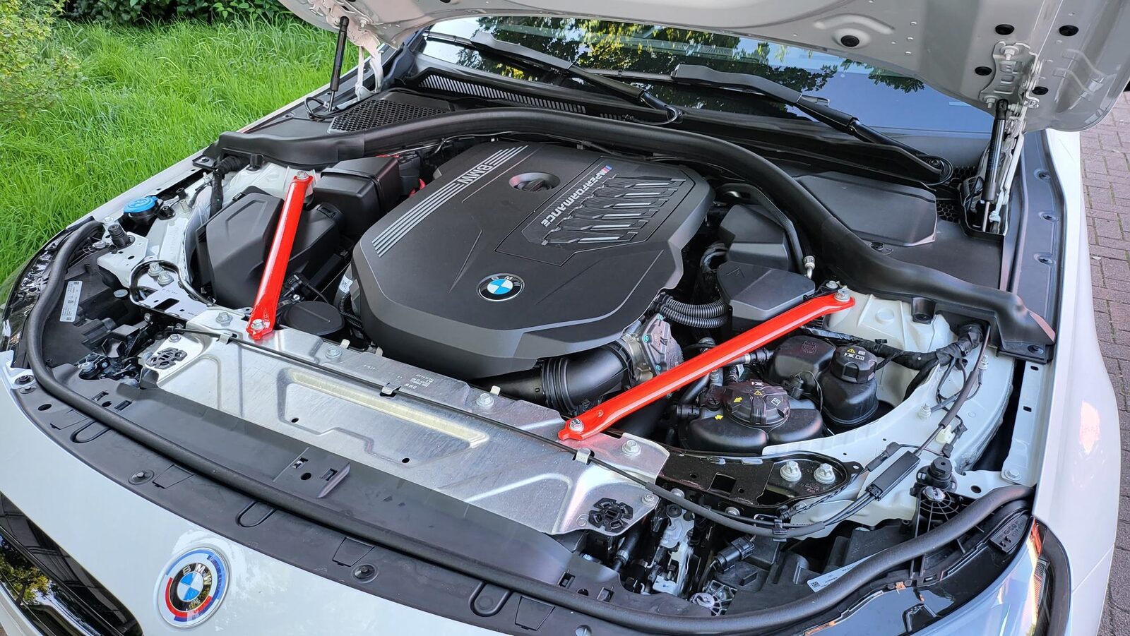 Öppet motorrum i en vit BMW M240i med B58-motorn och två röda fjäderbensstag, fotograferat utomhus med gräsmatta i bakgrunden