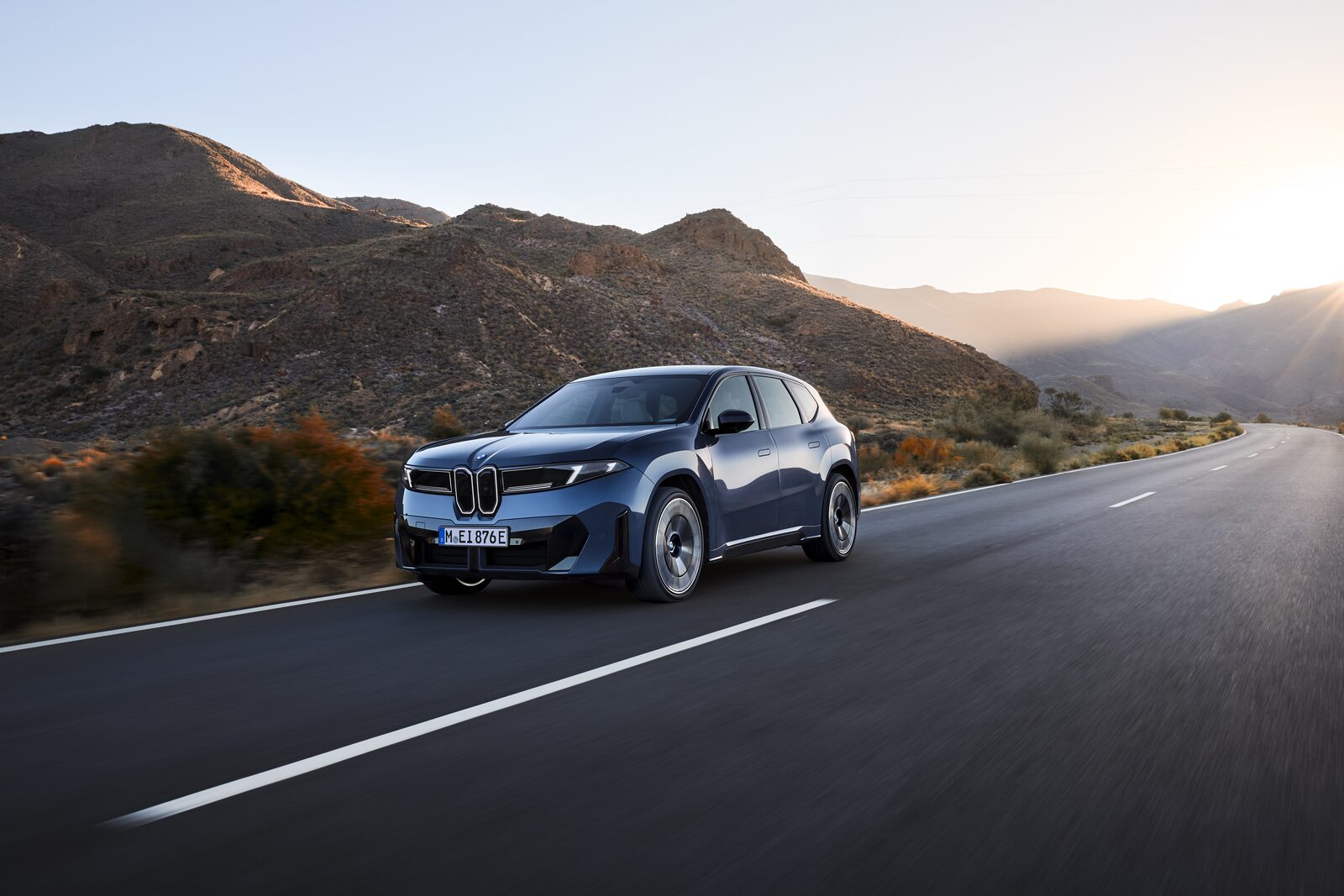 Blå BMW iX3 kör på en landsväg genom ett torrt bergslandskap i motljus
