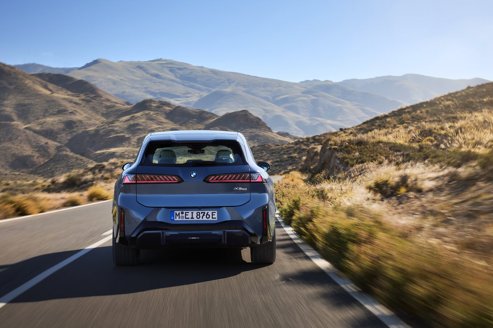 BMW iX3 bakifrån på en solig bergsväg med kullar och torrt gräs vid vägkanten