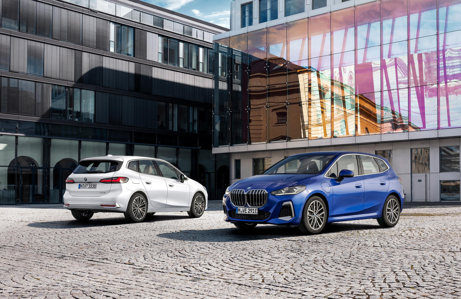 En vit och en blå BMW 2-serie Active Tourer parkerade på kullersten framför moderna glasfasader