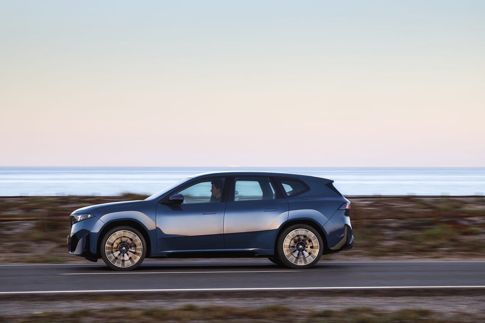 Gråblå BMW iX3 50 xDrive i fart längs en kustväg, sedd från sidan
