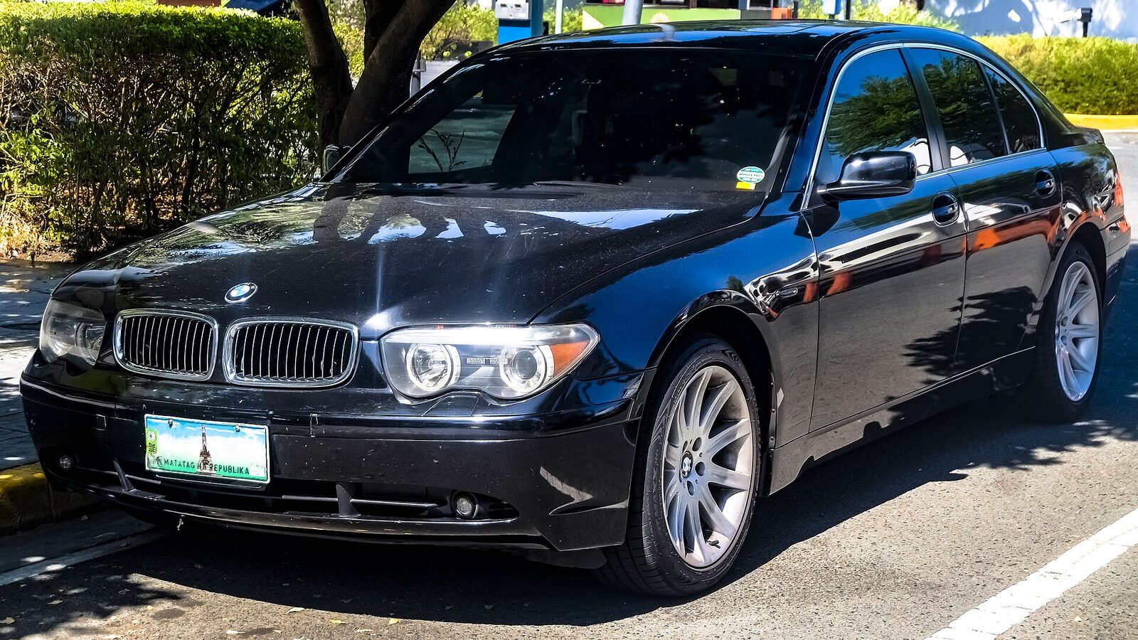Svart BMW 745i E65 snett framifrån, parkerad i skuggan under träd, med de karakteristiska ögonbrynsstrålkastarna och de breda njurarna