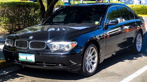 Svart BMW 745i E65 snett framifr&aring;n i skuggan under tr&auml;d, med &ouml;gonbrynsstr&aring;lkastare och breda njurar